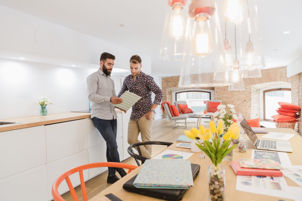 Two men stand by a kitchen counter looking at documents in a modern, bright office with laptops and flowers on the table.